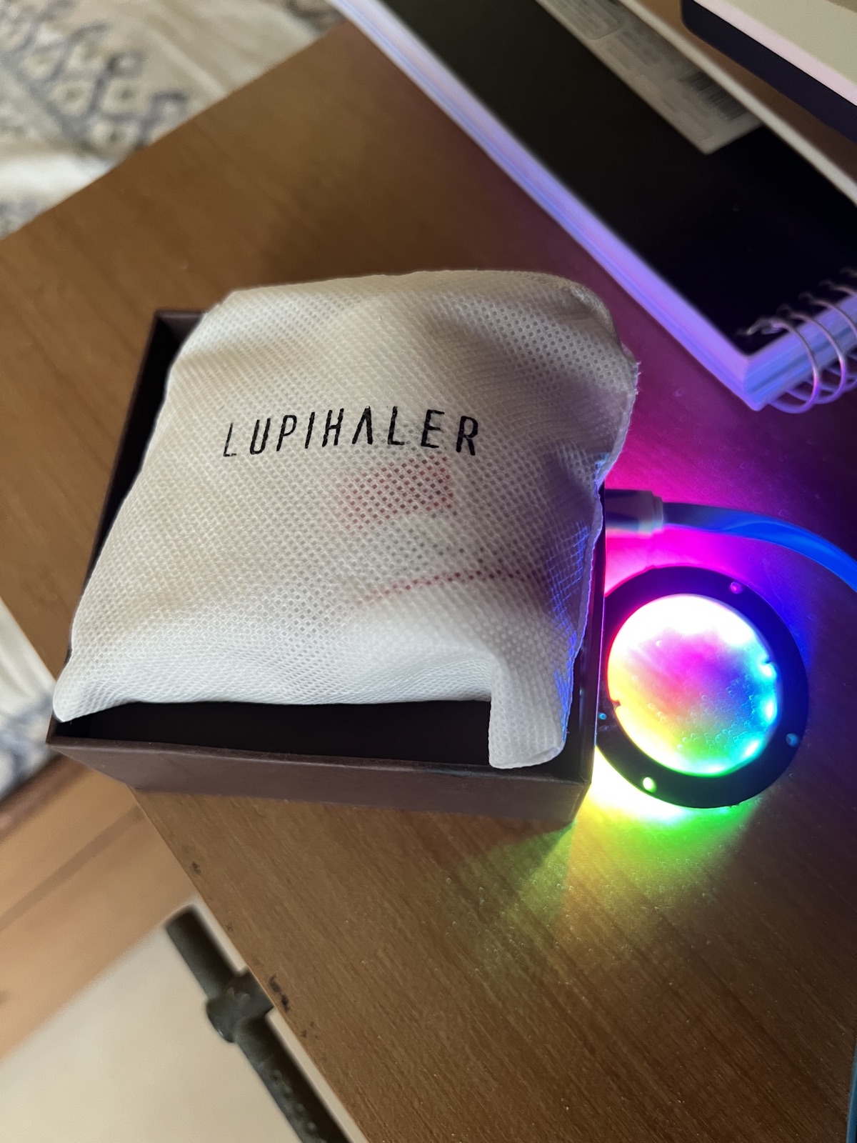 The Lumi prototype — a ring of light glowing in full rainbow colour beside an asthma inhaler in its pouch on a wooden desk.